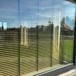 Zewnętrzne żaluzje fasadowe z czarnymi prowadnicami zamontowane w oknie, widoczne odbicie ogrodu i nieba na szkle.