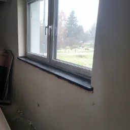 Montaż czarnego parapetu w surowym stanie, widok z boku na okno z widokiem na zewnątrz, ściany wymagają wykończenia.