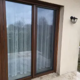 Przesuwne okno tarasowe z brązowymi ramami i moskitierą w jednej części, widoczne krople deszczu na szybie, jasna elewacja budynku i lampa zewnętrzna.