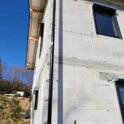 Surowy budynek z betonu komórkowego z zamontowanymi oknami i czarną rurą spustową, widoczne przygotowania pod montaż osłon okiennych.