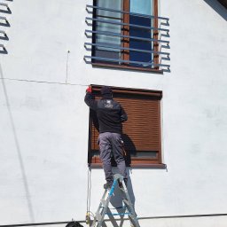 Letyro Bartosz Lekki - Pracownik na drabinie montuje brązowe osłony okienne na białej fasadzie domu, widoczne okna z ozdobnymi metalowymi kratami.