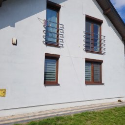 Letyro Bartosz Lekki - Biały dom jednorodzinny z brązowymi ramami okiennymi, na górnych oknach widoczne metalowe barierki, na dolnych oknach zamontowane rolety dzień noc, na ścianie widoczna skrzynka gazowa.