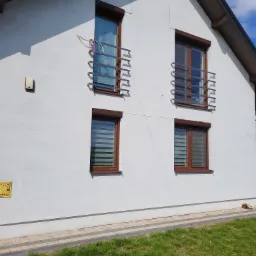 Biały dom jednorodzinny z brązowymi ramami okiennymi, na górnych oknach widoczne metalowe barierki, na dolnych oknach zamontowane rolety dzień noc, na ścianie widoczna skrzynka gazowa.