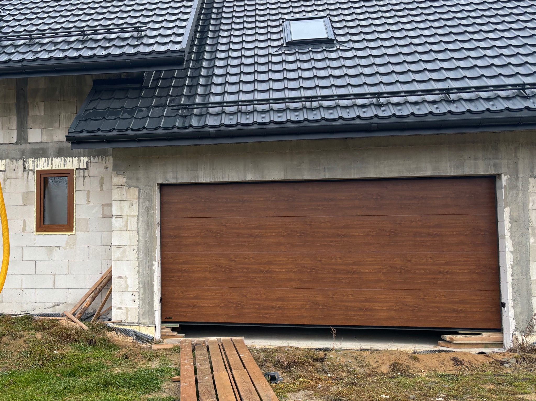 Zamknięta brama garażowa segmentowa w kolorze drewna, zamontowana w surowym budynku z białego bloczka, widoczne drewniane deski przed bramą.