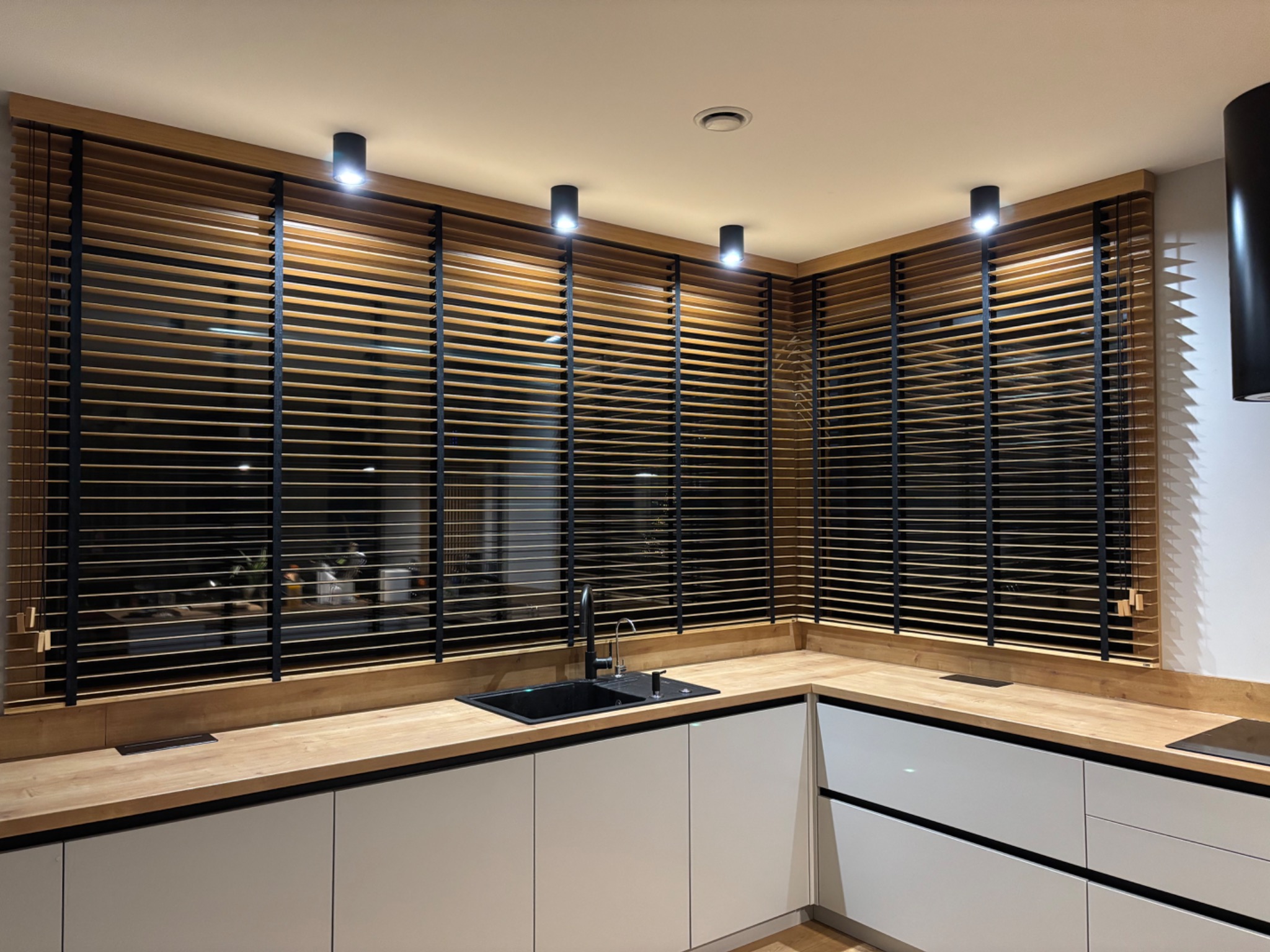 Drewniane żaluzje poziome w kolorze naturalnego drewna z czarnymi elementami, zamontowane w oknach nowoczesnej kuchni z białymi szafkami i drewnianym blatem.