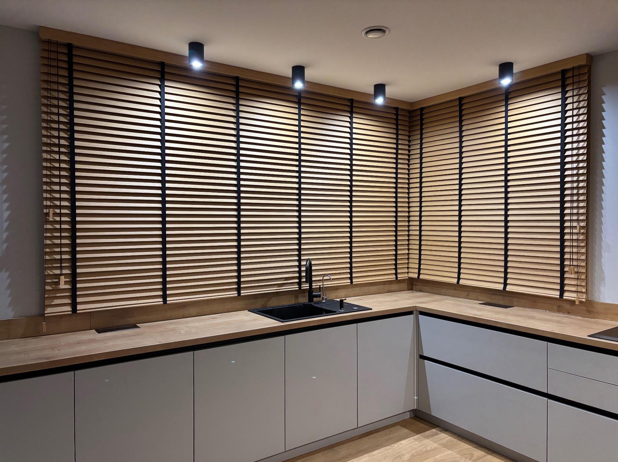 Jasne, drewniane żaluzje poziome z czarnymi taśmami w nowoczesnej kuchni z drewnianym blatem i szarymi szafkami, oświetlone punktowymi lampami sufitowymi.