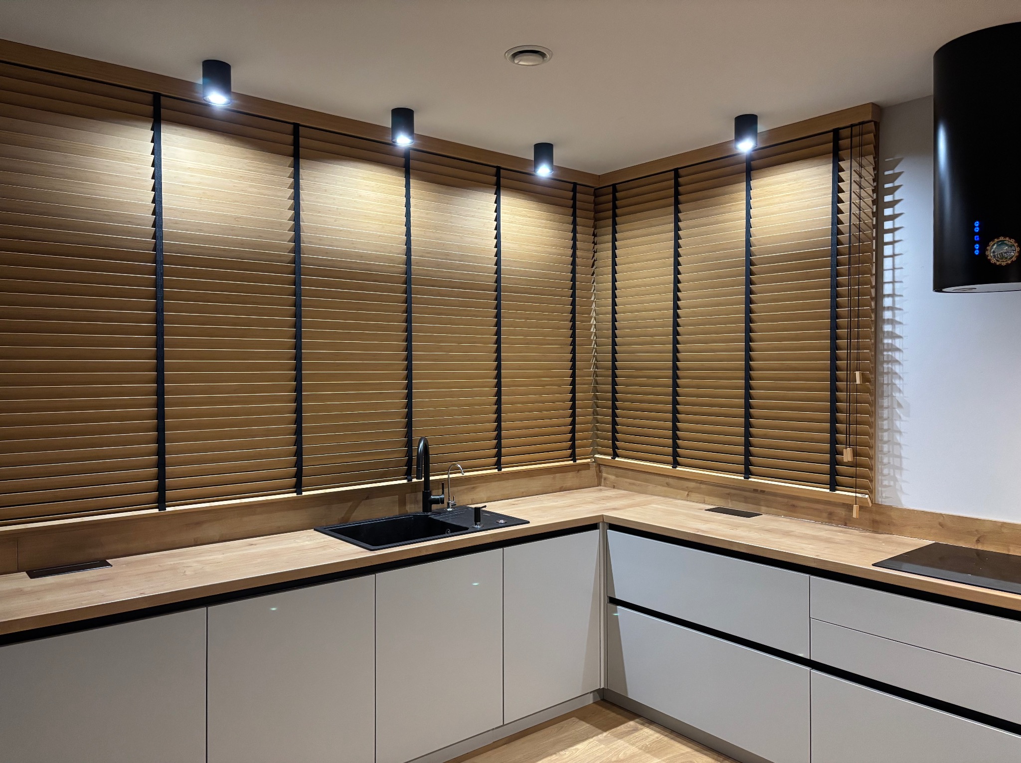 Drewniane żaluzje poziome z czarnymi taśmami w nowoczesnej kuchni z białymi szafkami i drewnianym blatem, oświetlone punktowymi lampami sufitowymi.