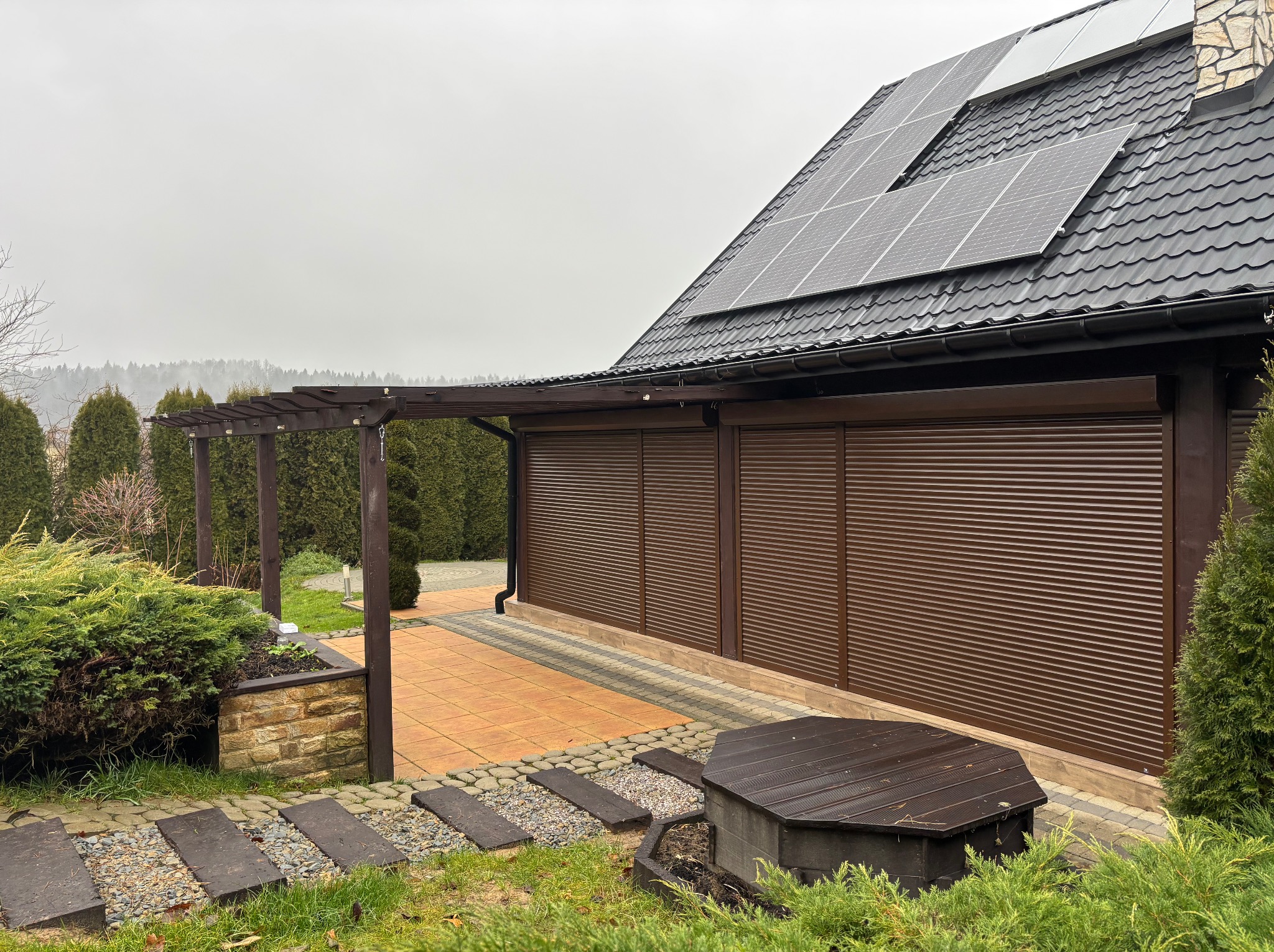 Brązowe, poziome panele osłonowe zamknięte na budynku z panelami słonecznymi na dachu, widok z ogrodu z pergolą i ścieżką.