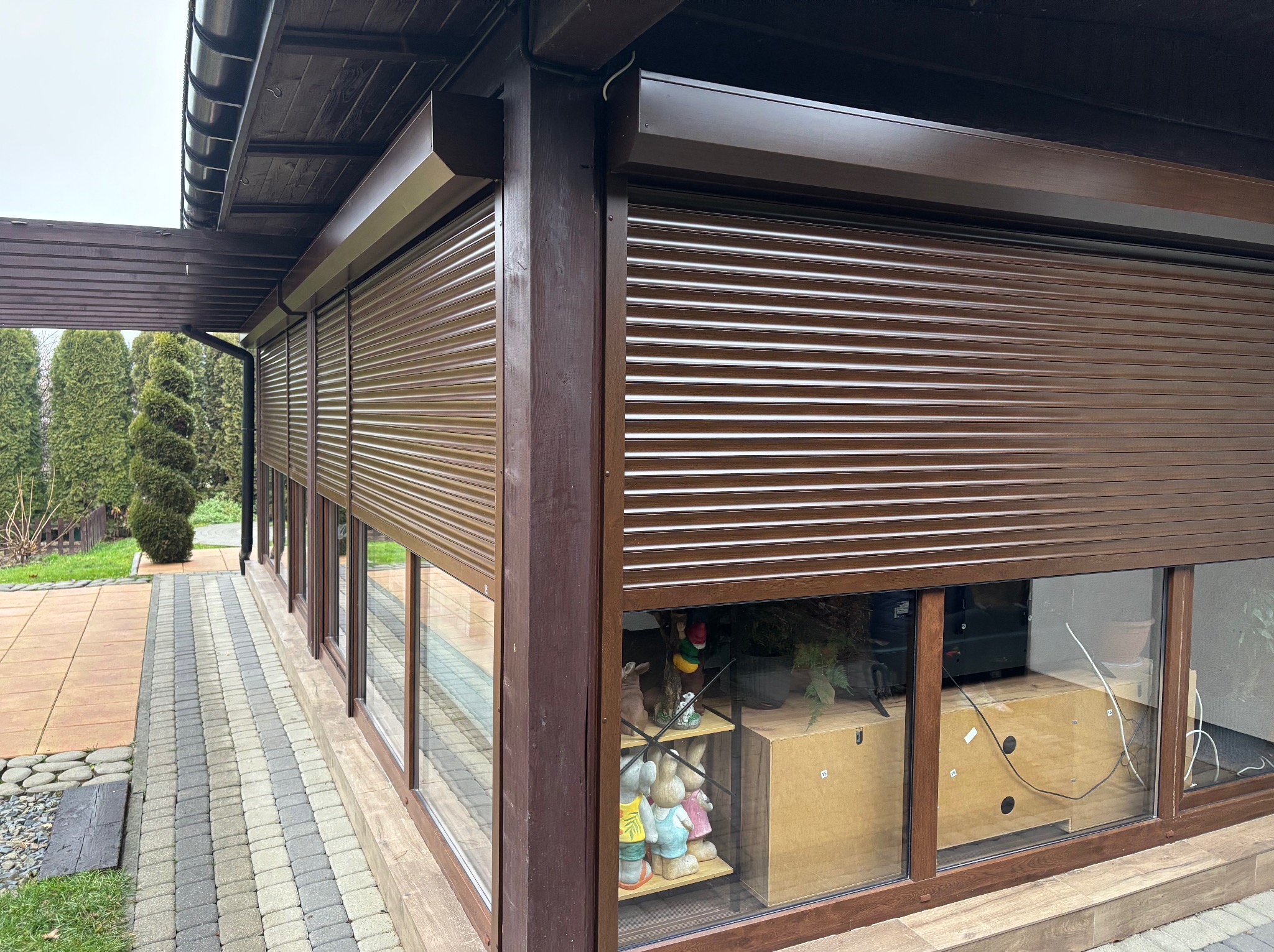 Zewnętrzne, brązowe rolety okienne całkowicie zasłonięte, zamontowane w drewnianej altanie z kostką brukową na zewnątrz.