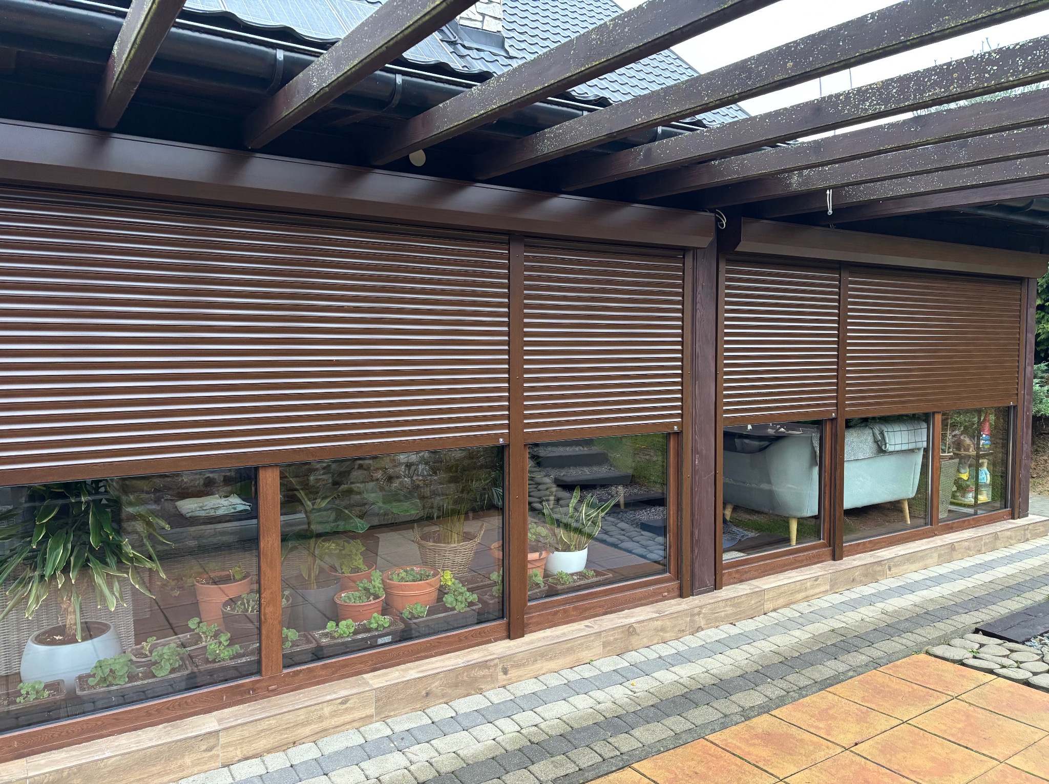 Zewnętrzne brązowe osłony okienne częściowo zasłonięte, widoczne rośliny doniczkowe w pomieszczeniu i na zewnątrz, drewniana pergola nad konstrukcją.