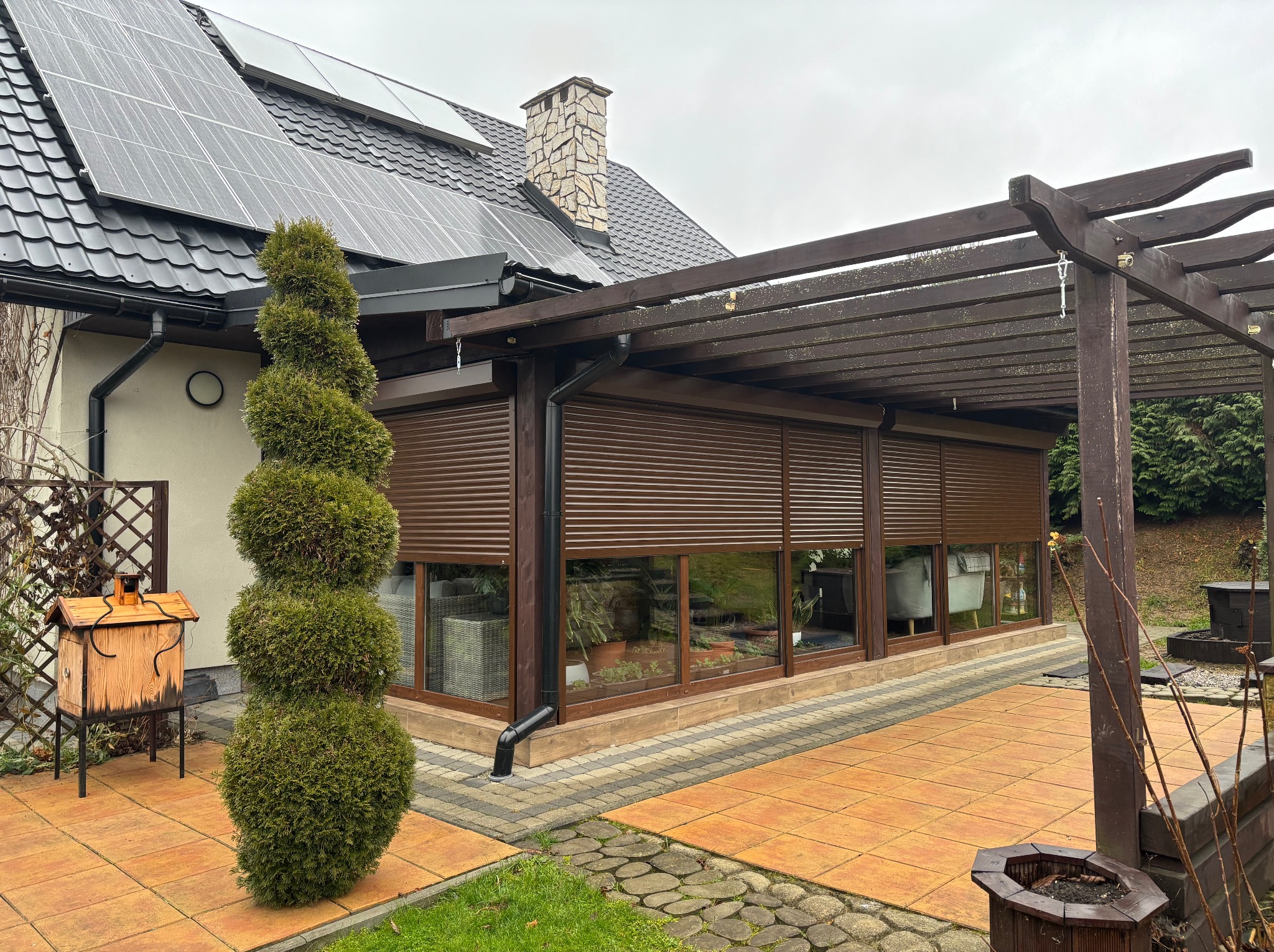 Zadaszony taras z brązowymi roletami zewnętrznymi, widok na ogród, na dachu panele słoneczne i komin z kamienia.
