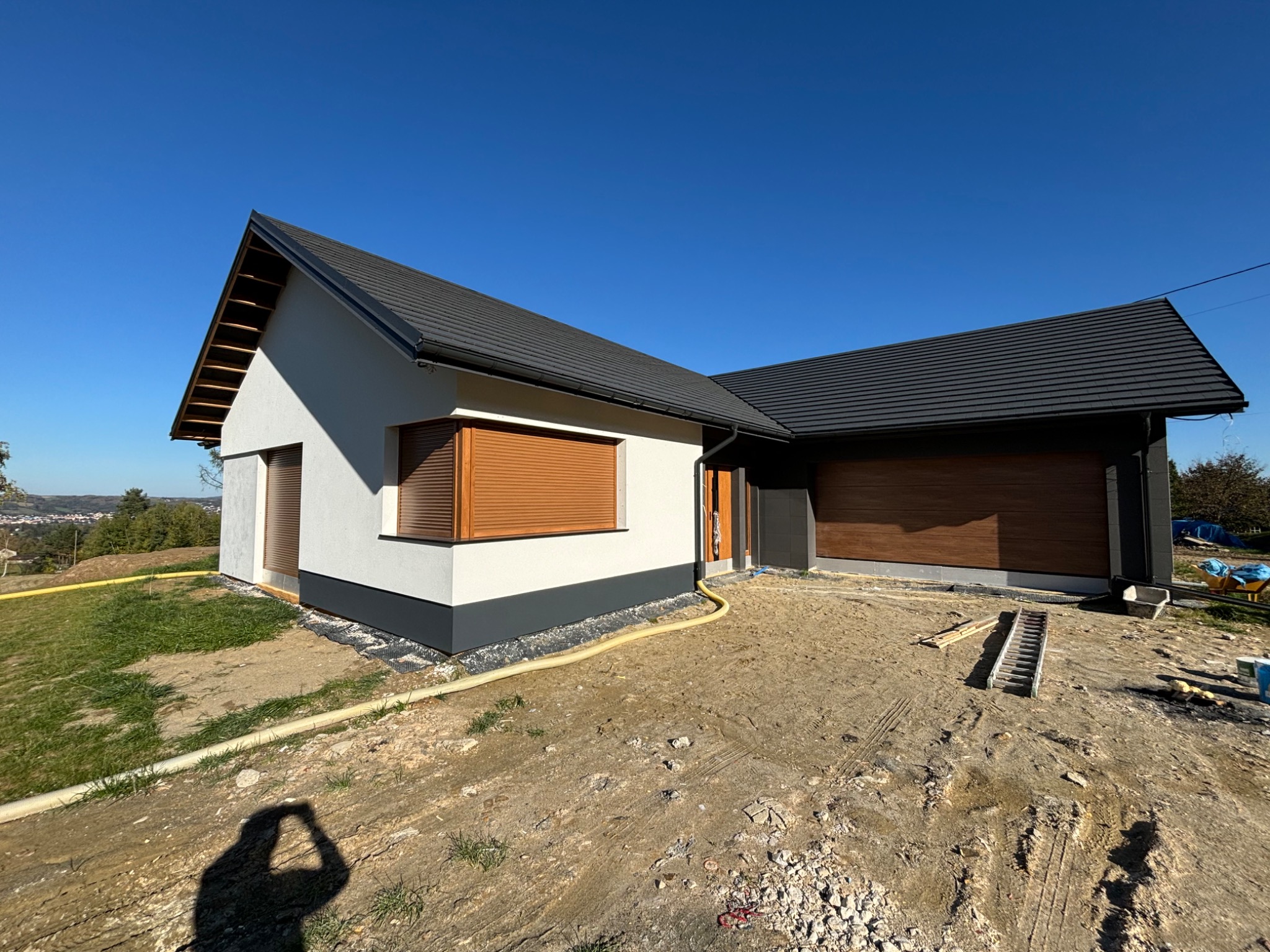 Nowoczesny dom jednorodzinny z grafitowym dachem i brązowymi roletami zewnętrznymi, widok z zewnątrz na placu budowy, cień fotografa w lewym dolnym rogu.