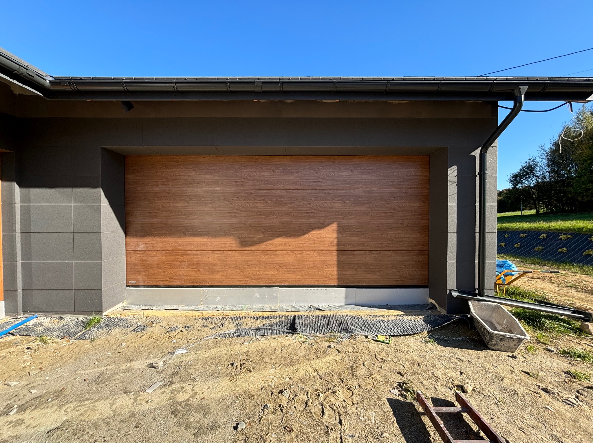 Nowa, segmentowa brama garażowa z drewnopodobnym panelem, zamontowana w nowoczesnym domu z szarą elewacją, widoczna w trakcie prac wykończeniowych na zewnątrz.