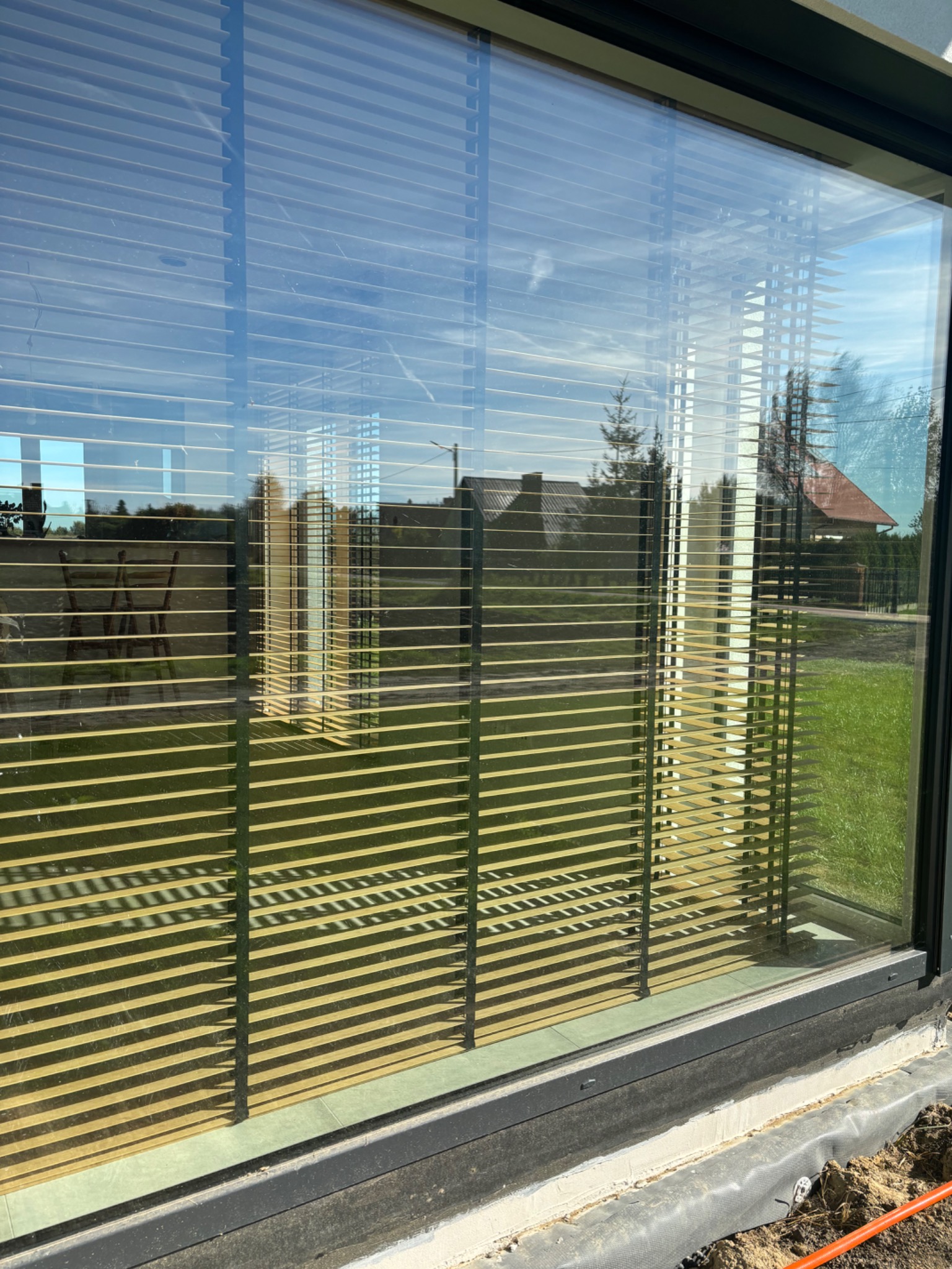 Zewnętrzne żaluzje fasadowe z czarnymi prowadnicami zamontowane w oknie, widoczne odbicie ogrodu i nieba na szkle.