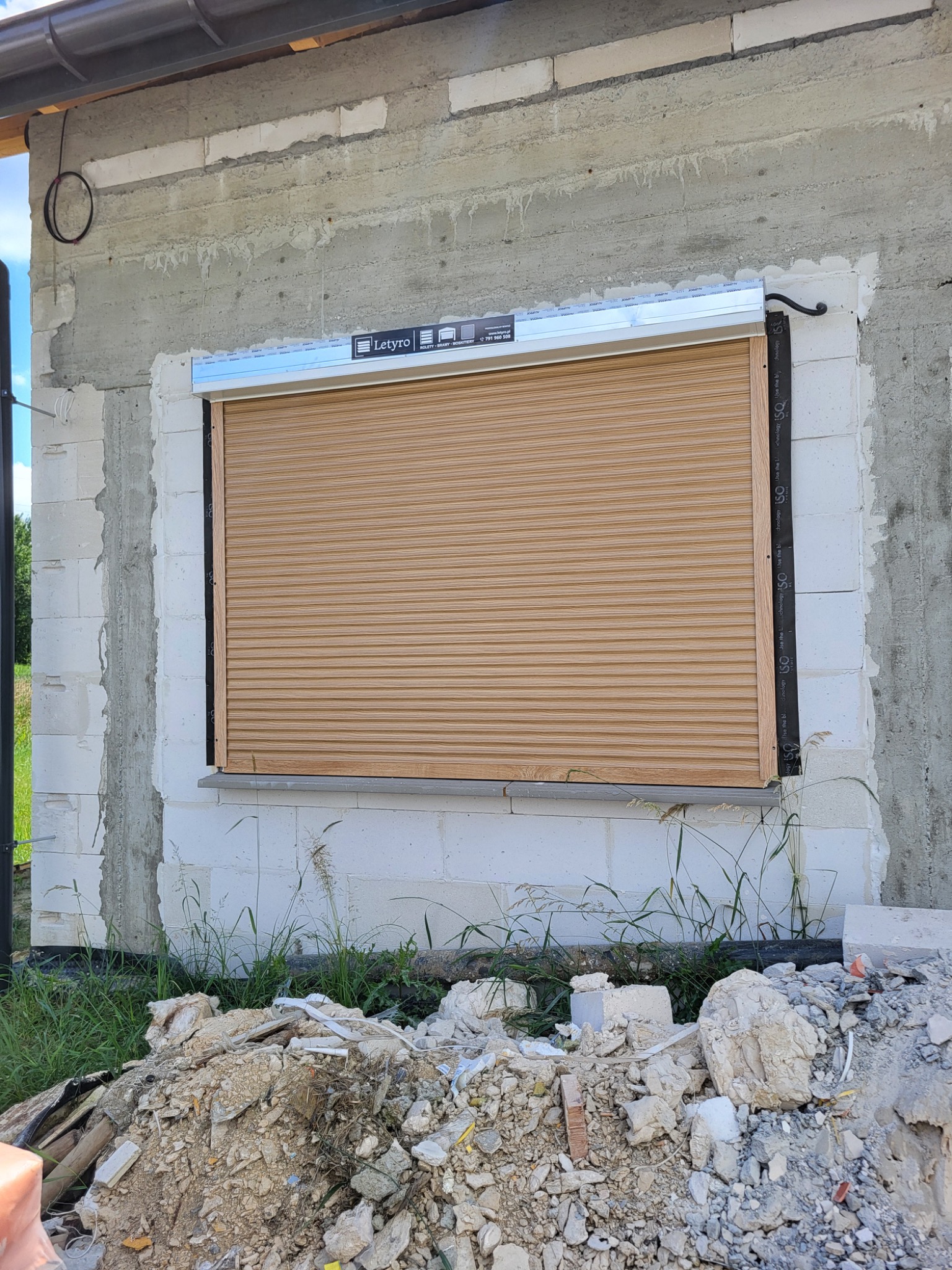 Zamknięta, drewnopodobna roleta zewnętrzna w oknie surowego budynku z betonu komórkowego, widoczne ślady montażu i gruz przed oknem.