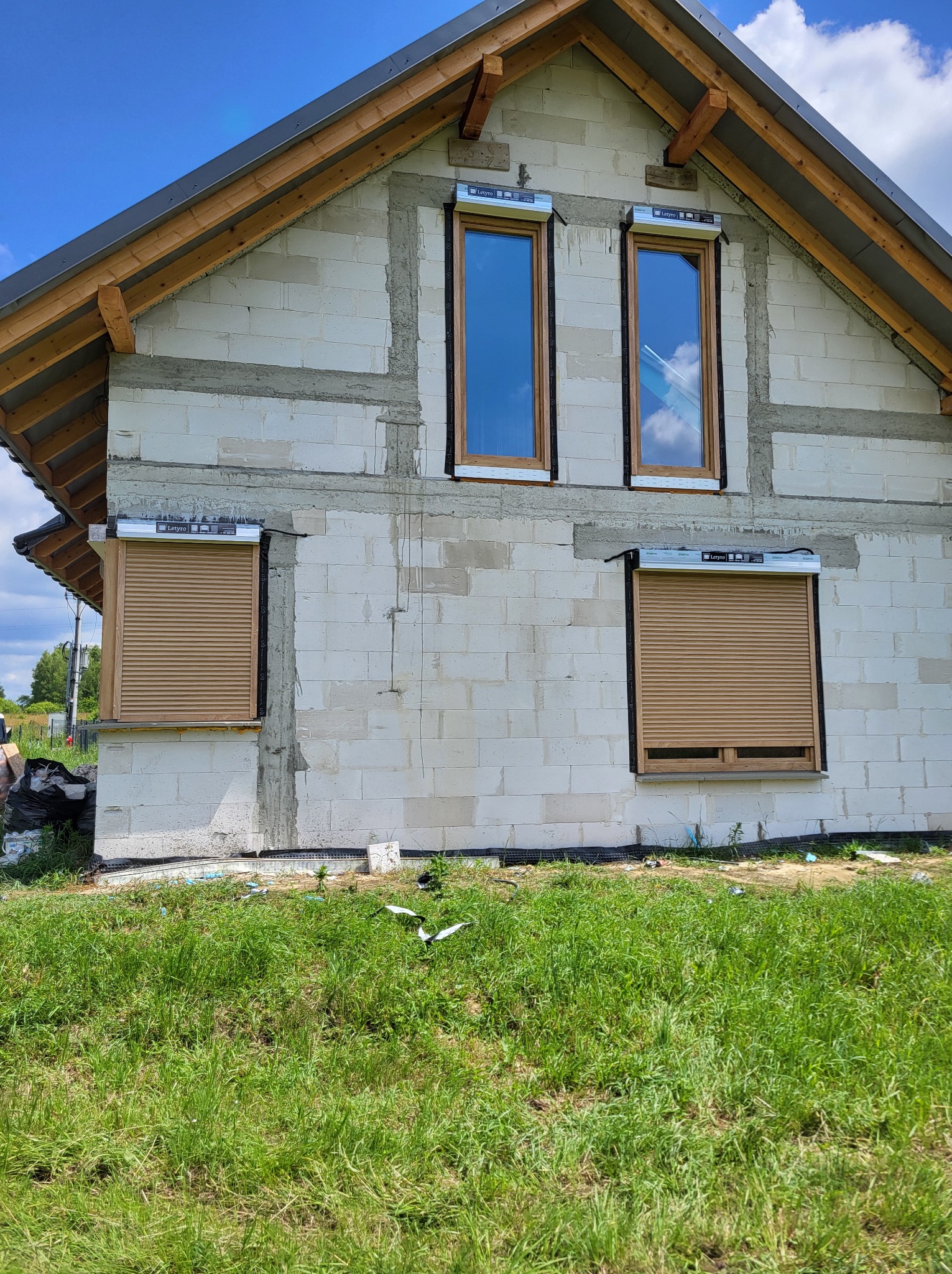 Dom w trakcie budowy z zamontowanymi roletami zewnętrznymi w kolorze drewna na parterze, okna na piętrze jeszcze bez rolet, widoczne materiały budowlane i trawa.