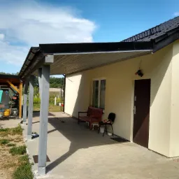 Dom z nowym dachem z czarnej dachówki i świeżo pomalowaną elewacją w odcieniach beżu, widok z zewnątrz na zadaszony taras z ławką i rowerem w tle.