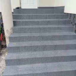 Schody wejściowe wyłożone szarą, ziarnistą płytką ceramiczną, prowadzące do białych drzwi wejściowych, z widocznym fragmentem kolumny z odpryskującym tynkiem po lewej stronie.