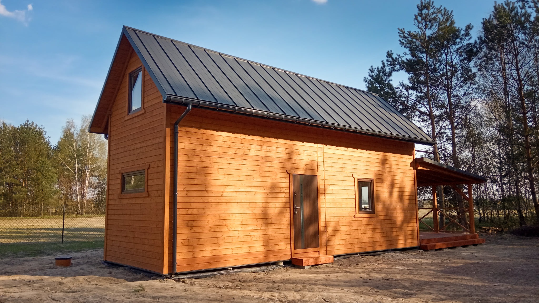 Nowoczesny, parterowy dom z drewnianą elewacją, ciemnym dachem i małym zadaszonym tarasem, otoczony drzewami.