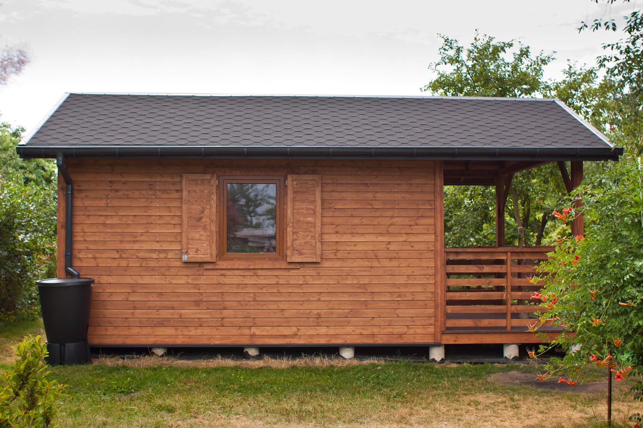 Drewniany domek z brązowym dachem, widoczną werandą i zbiornikiem na deszczówkę, otoczony zielenią.