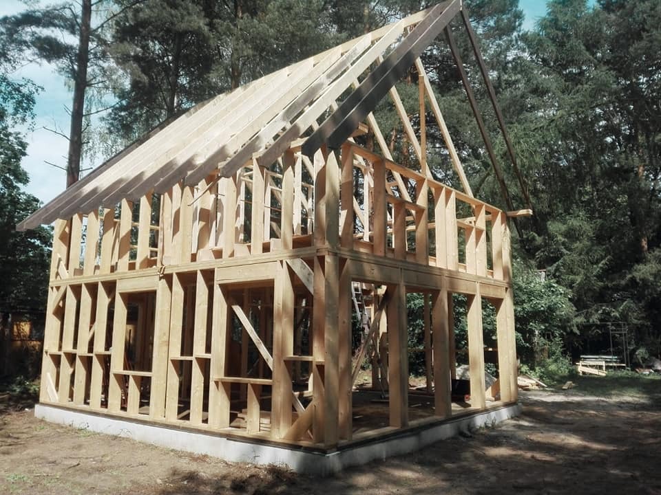 Surowy szkielet drewnianego domu w trakcie budowy, widoczne belki i krokwie, w tle las iglasty, fundament betonowy.