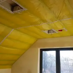 Żółta folia paroizolacyjna szczelnie pokrywa skośny sufit poddasza, zabezpieczona białymi listwami, dwa otwory na oświetlenie punktowe, widok na okno dachowe i drzewa na zewnątrz.