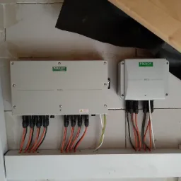 Dwa szare moduły sterujące z logo PROJOY, zamontowane na ścianie z białych bloczków, z widocznymi podłączonymi przewodami w kolorze pomarańczowym, czarnym, białym i żółto-zielonym.