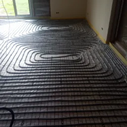 Rozłożona instalacja ogrzewania podłogowego na folii z siatką, rury ułożone spiralnie, widoczne okna i przygotowane ściany, poziomica leży na rurach.