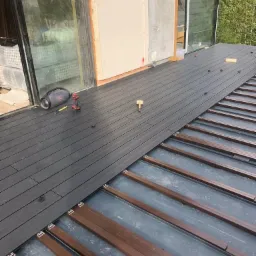 Montaż deski kompozytowej na tarasie: widoczne legary, częściowo ułożona podłoga, w tle okno tarasowe i fragment elewacji budynku.