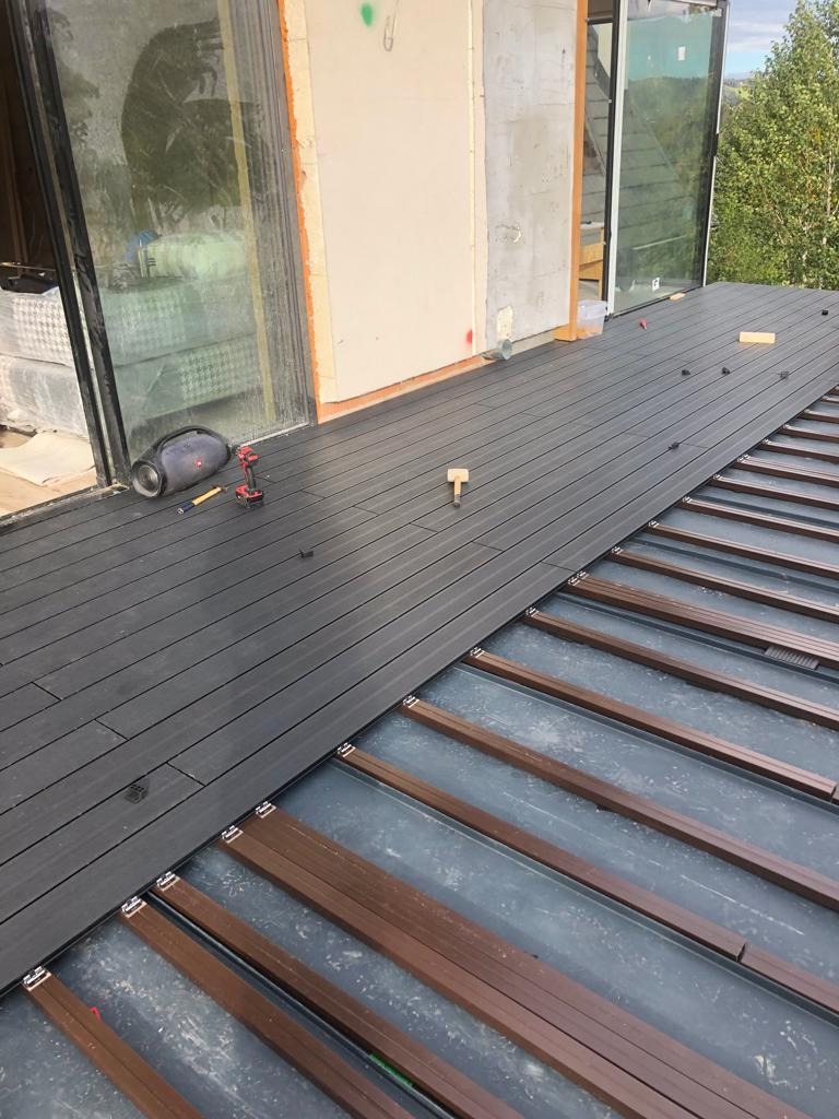 Montaż deski kompozytowej na tarasie: widoczne brązowe legary, częściowo ułożona szara deska, w tle okno tarasowe i ściana budynku w trakcie wykończenia, w pobliżu wiertarka akumulatorowa i głośnik.