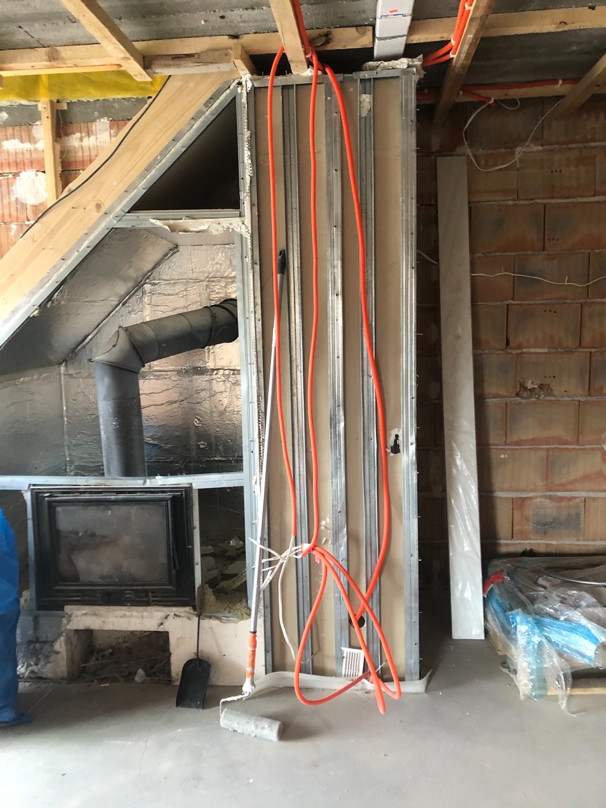 Wnętrze w trakcie prac przygotowawczych: widoczne profile metalowe, pomarańczowe rury instalacyjne, kominek z obudową w trakcie tworzenia, częściowo ocieplony, oraz narzędzia malarskie leżące...