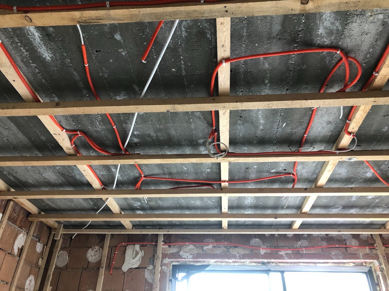 Instalacja elektryczna w pomarańczowych rurkach ochronnych, prowadzona pod surowym, betonowym stropem, zamocowana do drewnianej konstrukcji szkieletowej w trakcie prac wykończeniowych wnętrza.