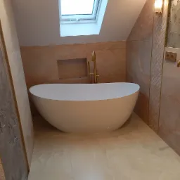 Nowoczesna, wolnostojąca wanna w beżowej łazience na poddaszu z oknem dachowym, armatura w kolorze złotym.