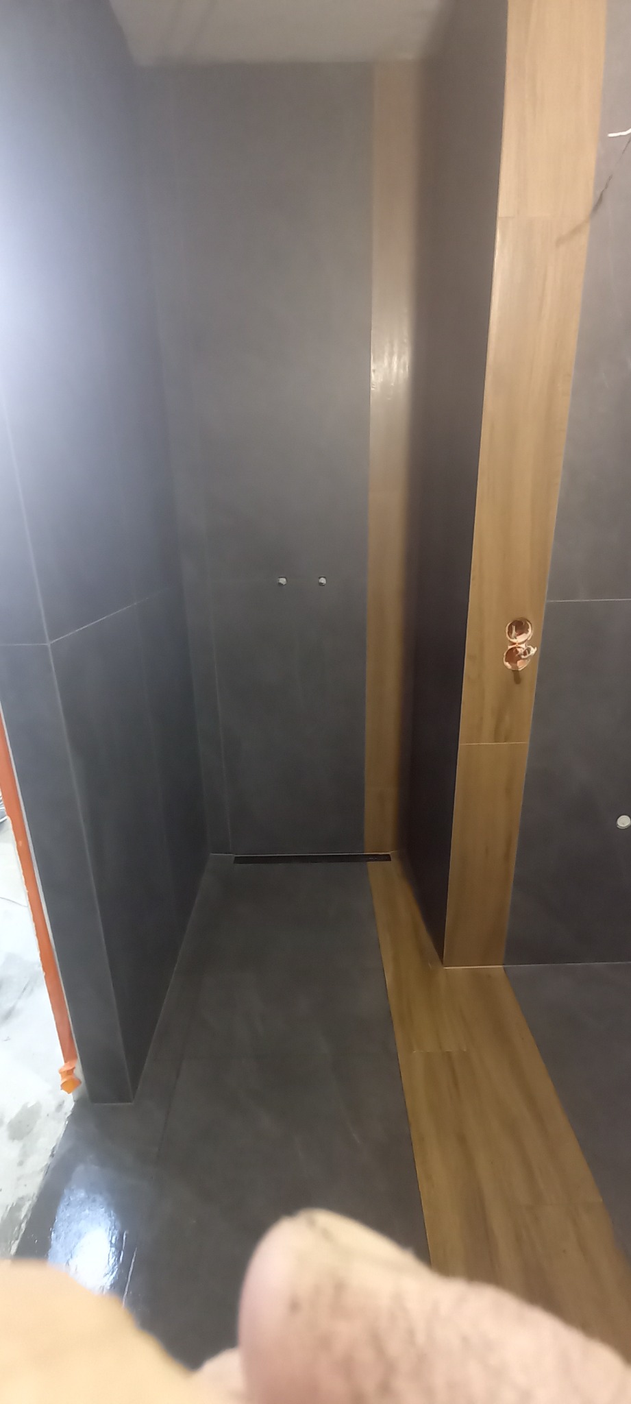 Wnętrze nowej kabiny prysznicowej z szarymi płytkami na ścianach i podłodze oraz drewnopodobnymi płytkami na fragmencie ściany, widoczny odpływ liniowy w podłodze i wyprowadzenia instalacji wodnej...