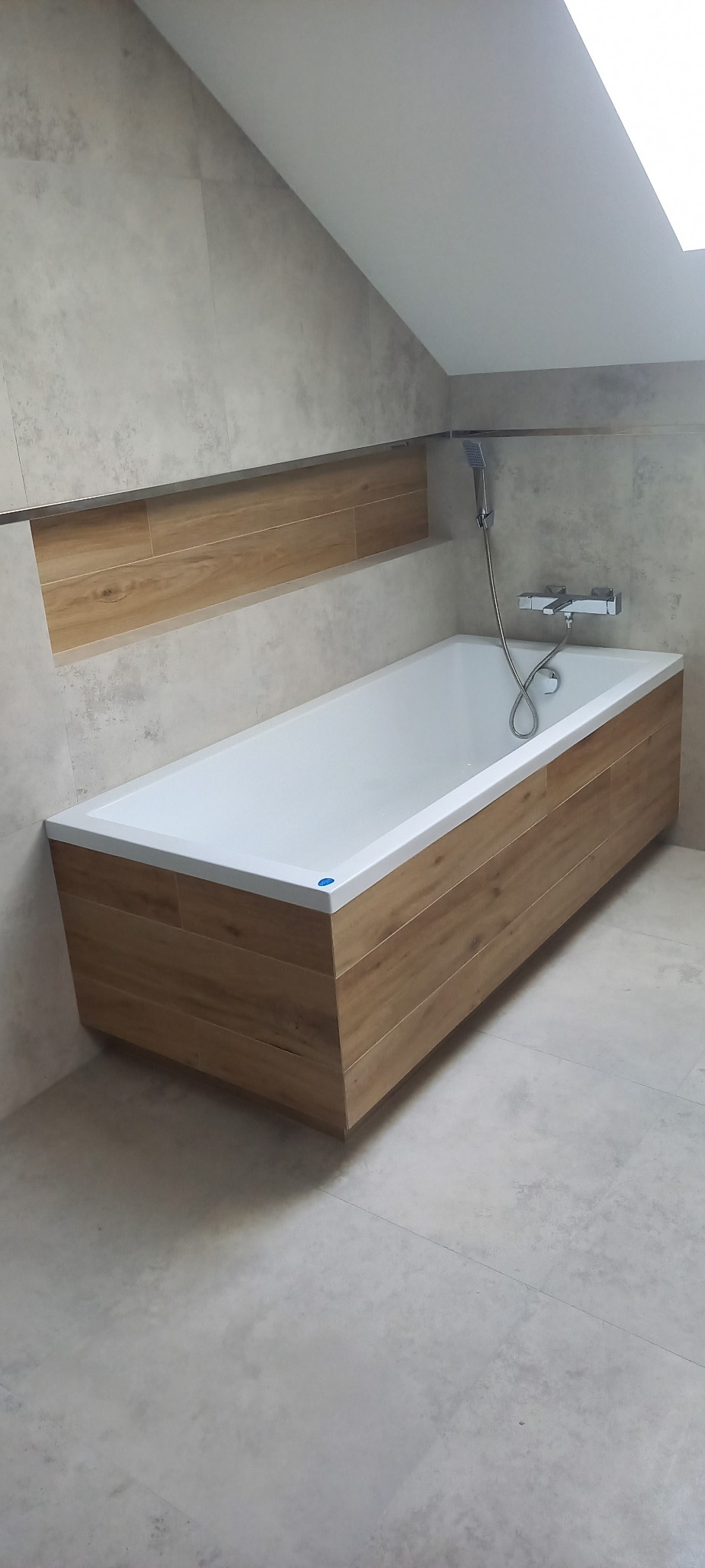 Nowoczesna wanna obudowana panelami imitującymi drewno, z chromowaną armaturą i prysznicem ręcznym w łazience wykończonej płytkami w odcieniach szarości, oświetlonej światłem dziennym z okna...