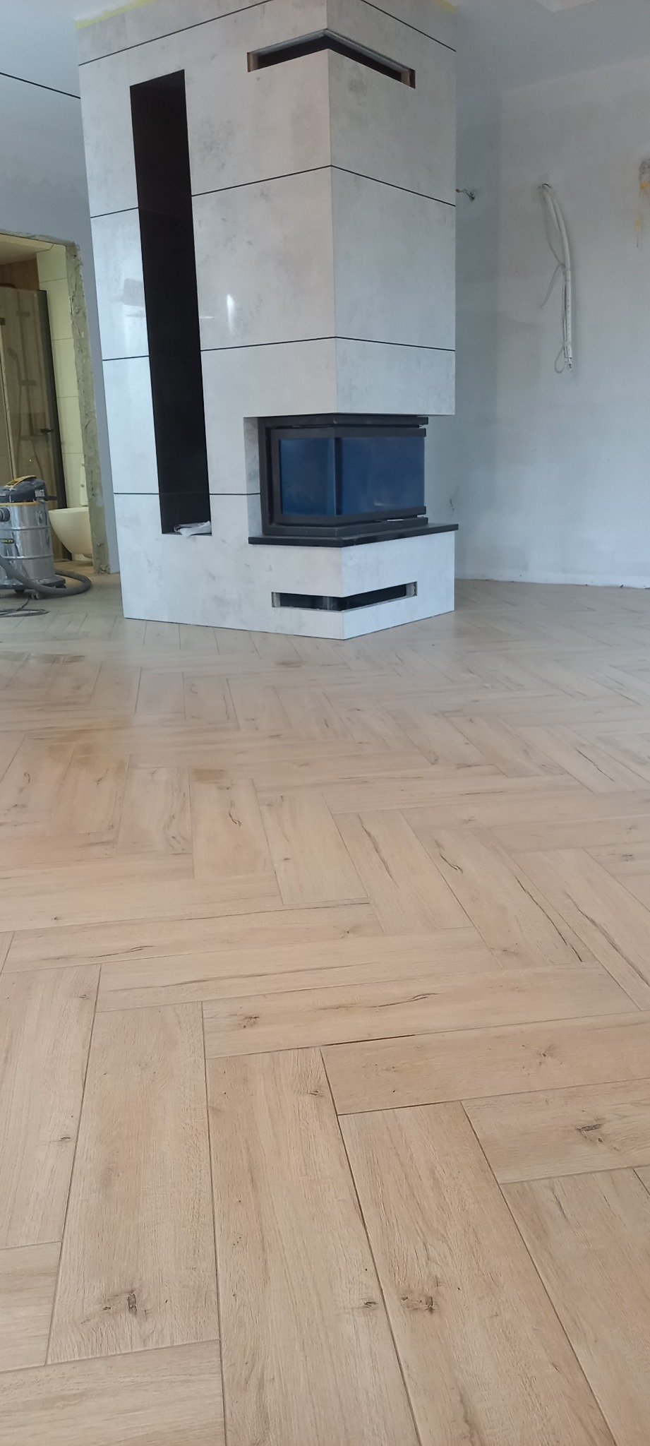 Częstochowa: Nowoczesny kominek obłożony płytkami w jasnych odcieniach szarości, z czarnym wkładem, na podłodze ułożonej w jodełkę z paneli imitujących drewno, widoczny odkurzacz przemysłowy...