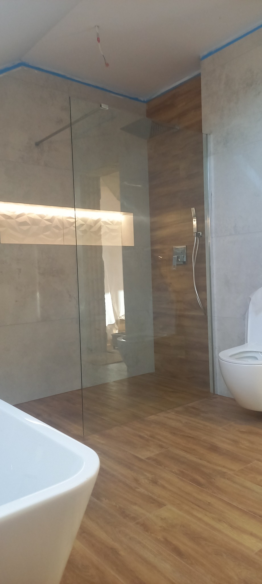 Nowoczesna łazienka z drewnopodobną podłogą, kabiną prysznicową typu walk-in, podświetlaną wnęką z dekoracyjną płytką 3D i wiszącą toaletą.