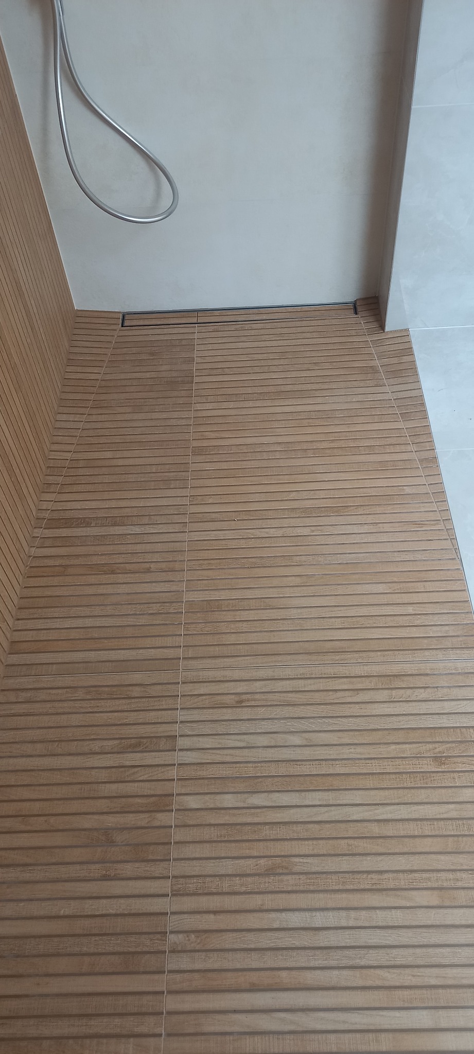 Ukończona podłoga i fragment ściany w kabinie prysznicowej, wyłożone płytkami imitującymi drewniane deski, z odpływem liniowym przy ścianie.