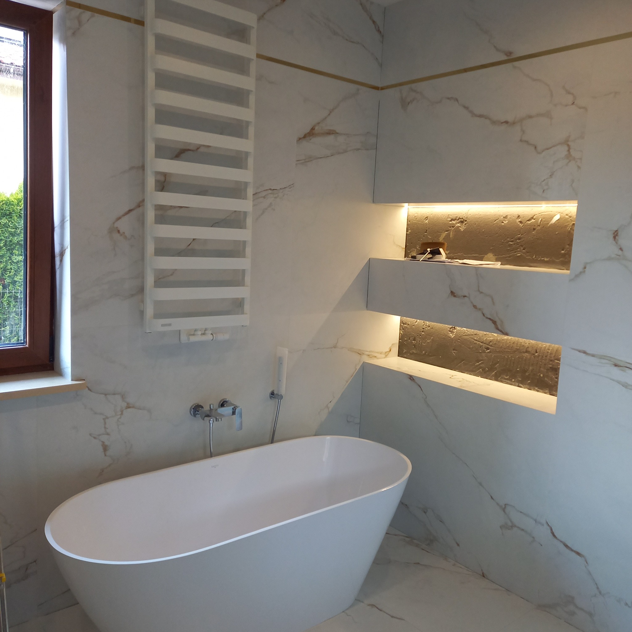 Nowoczesna łazienka z wolnostojącą białą wanną, grzejnikiem drabinkowym, marmurowymi płytkami z złotymi żyłami i podświetlanymi niszami w ścianie.