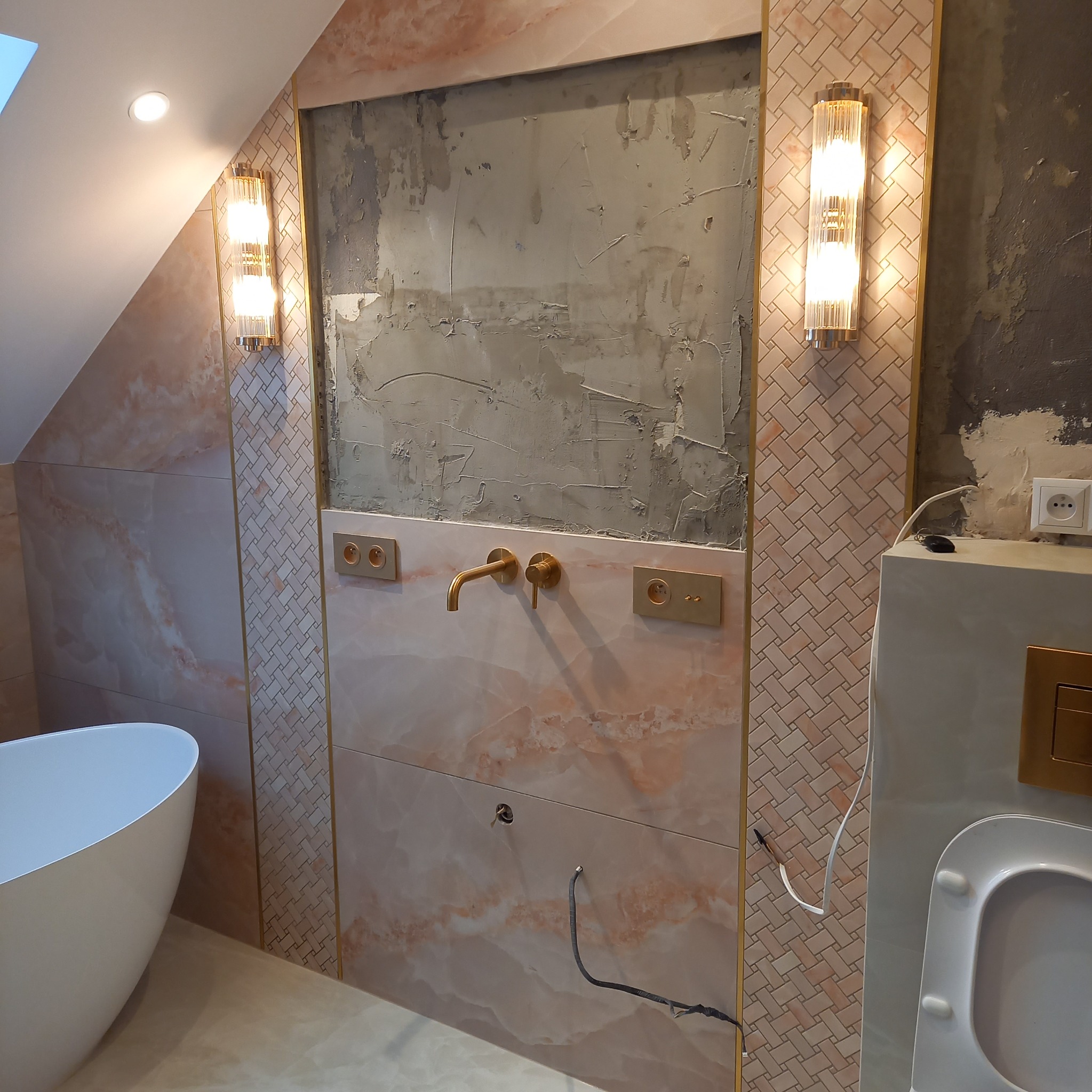 Niedokończona ściana w łazience z marmurowymi płytkami, złotą armaturą i ozdobnymi kinkietami, widoczne ślady po szpachlowaniu i wystające kable.