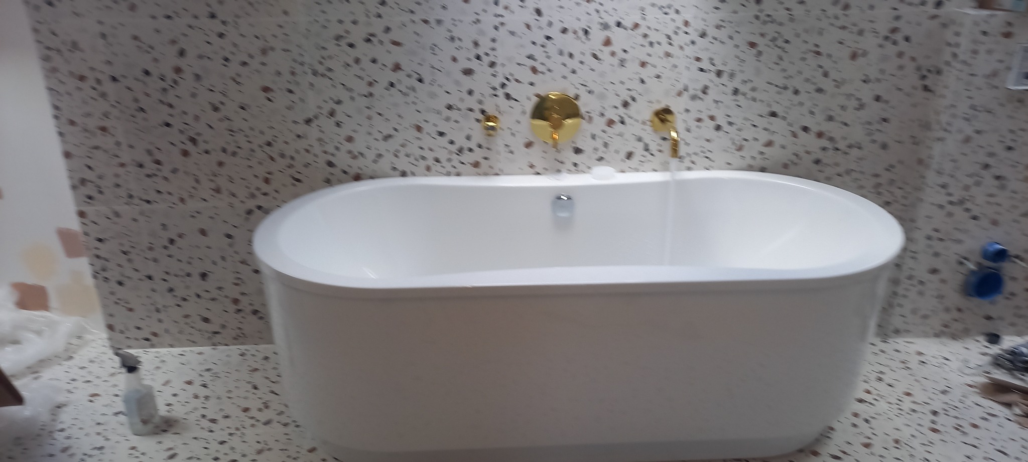 Biała wanna wolnostojąca z złotą armaturą na tle ściany i podłogi wyłożonej płytkami imitującymi lastryko.