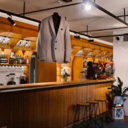 Jasnoszary męski garnitur wiszący na wieszaku nad drewnianym barem z butelkami i kieliszkami w tle, oświetlony punktowymi lampami.