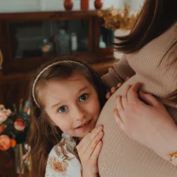 C&oacute;rka przytula się do brzucha ciężarnej mamy, w tle widoczny drewniany kredens i wazon z kwiatami.