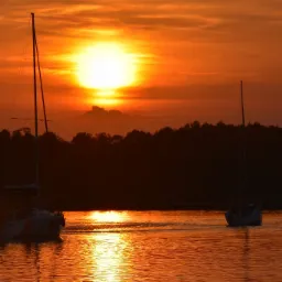 Dwa żaglowce płynące po wodzie w kierunku zachodzącego słońca, odbijającego się w tafli jeziora, z ciemnym zarysem lasu na horyzoncie.