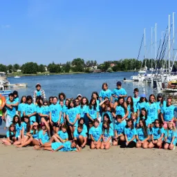 Duża grupa dzieci i młodzieży w błękitnych koszulkach z napisem 'AKTYWNE 2022' pozuje na piaszczystej plaży nad jeziorem, w tle zacumowane jachty z wysokimi masztami.