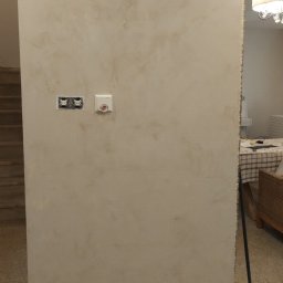 Titanhand - Ściana wewnętrzna z dekoracyjną, beżową strukturą, widoczne gniazdka elektryczne i czujnik, zabezpieczenia podłogi folią malarską, fragment schodów i mebli w tle.