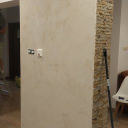 Titanhand - Ściana wewnętrzna po nałożeniu dekoracyjnej, beżowej farby strukturalnej, z widocznymi gniazdkami elektrycznymi i fragmentem kolumny wykończonej kamieniem dekoracyjnym. Na podłodze folia ochronna...