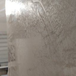 Titanhand - Ściana pokryta stiukiem weneckim Carmel, widoczne nieregularne wzory i faktura, delikatne refleksy światła, fragment białej szafki w tle.