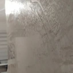 Ściana pokryta stiukiem weneckim Carmel, widoczne nieregularne wzory i faktura, delikatne refleksy światła, fragment białej szafki w tle.