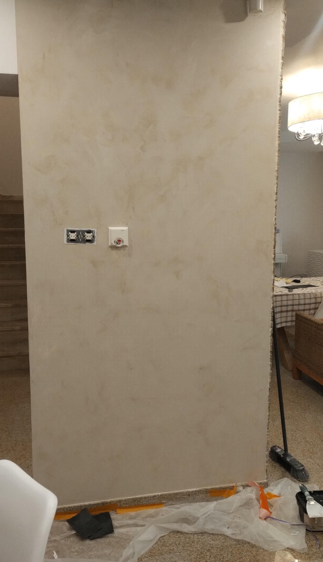 Ściana wewnętrzna z dekoracyjną, beżową strukturą, widoczne gniazdka elektryczne i czujnik, zabezpieczenia podłogi folią malarską, fragment schodów i mebli w tle.